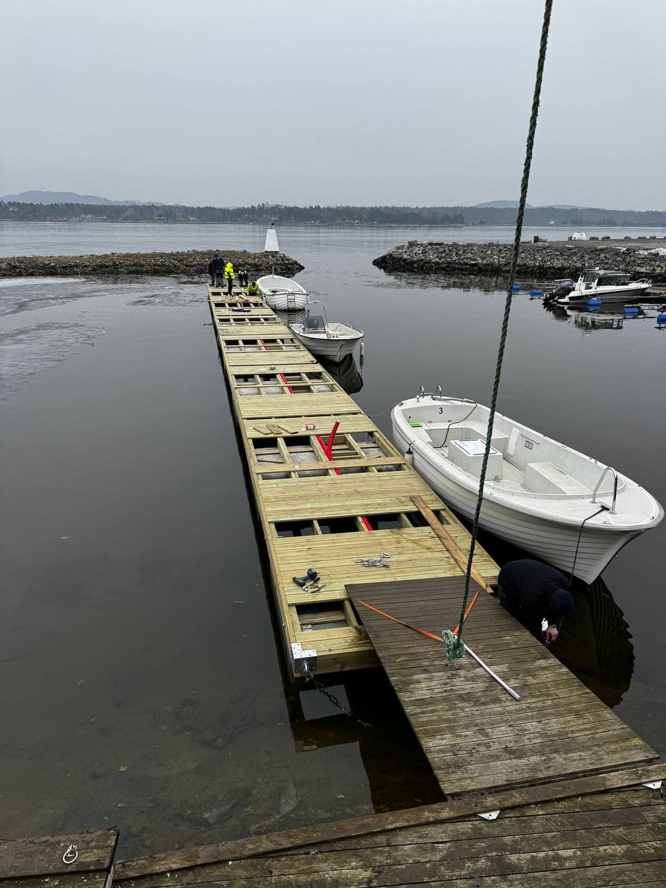 Flytebrygge under montering: bjelkelag i tre på betongelementer, med båter fortøyd langs siden.